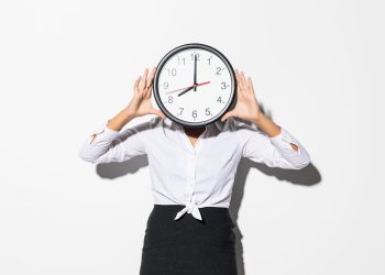 Photo of funny woman in white shirt and black skirt covering face with big round clock isolated over gray background
